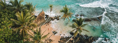 view from the sky of beautiful Beach and Palm trees with two People walking on beach and climbing on rocks