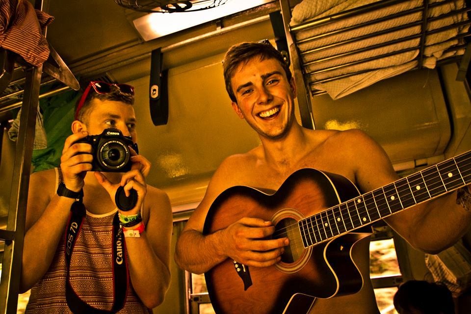 Two guys on a moving train, one with a camera taking photos and one with a guitar