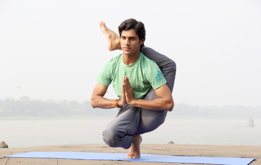 May doing flexible yoga with leg behind his back wearing green top and grey bottoms