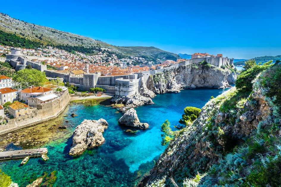 A aerial view of Dubrovnik, Croatia showing bright clear blue water, the historical sea walls and luscious greenery surrounding Croatia 