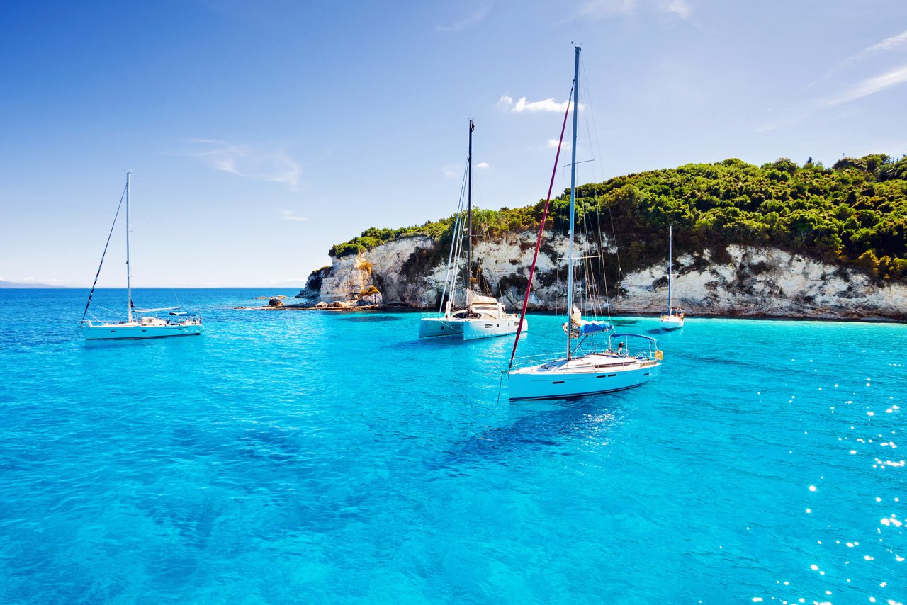 A view of three sailboats on the bright blue water in Greece with a background of white cliffs and greenery
