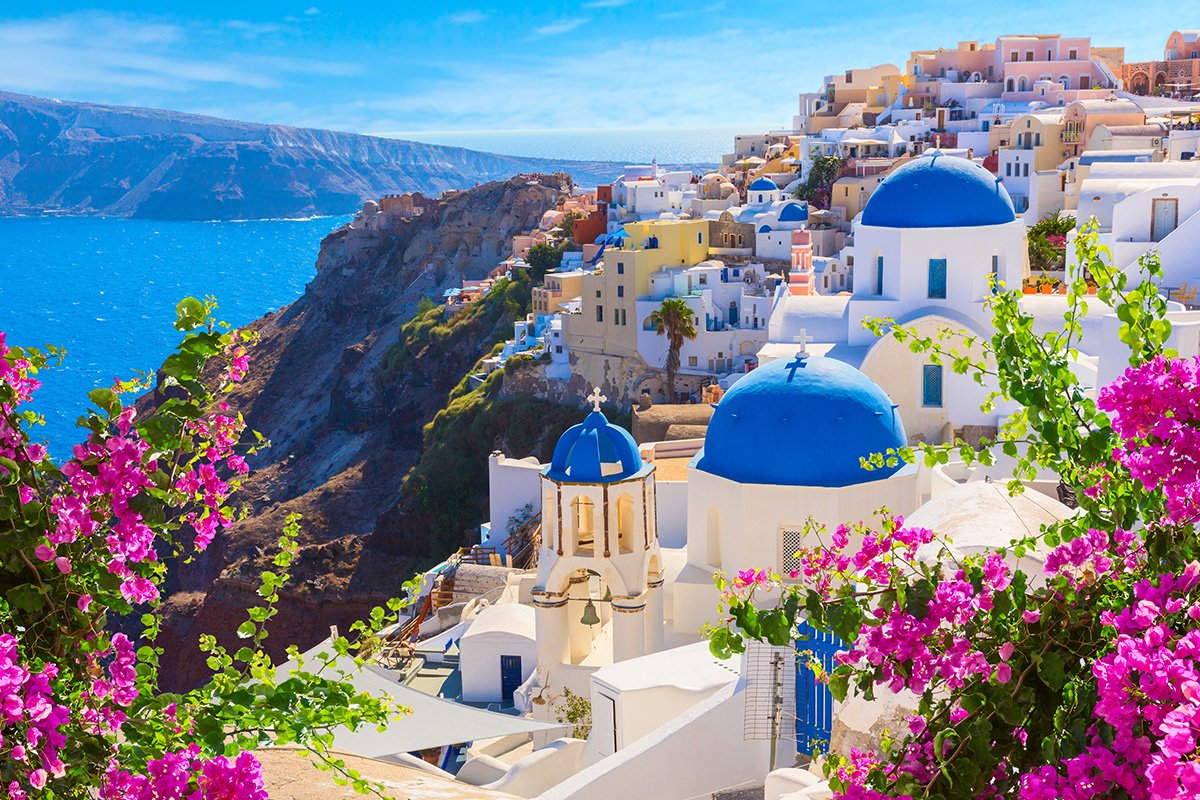 A scenic view of the white villas in Santorini from behind pink flowers with the mountains and sea in the background