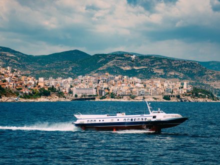 A ferry on the water in Greece with a view of the town and mountains in the background.