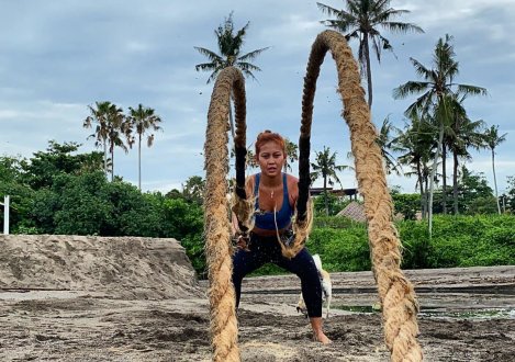 A girl using the battle ropes on the beach in Bali, Indonesia with a cloudy sky and palm trees in the background