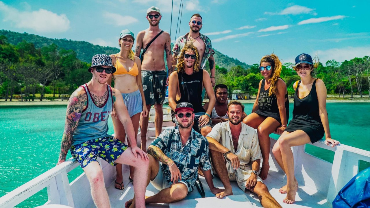Group of travelelrs sat on boat wearing colourful shirts, sea and blue sky in background
