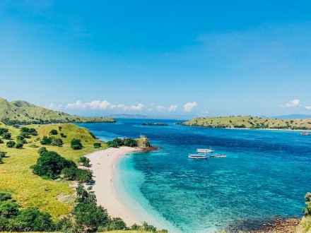 A scenic photo of a lush green landscape, white sandy beach and bright blue crystal clear water with the Komodo Island Hopper boat in the background in Indonesia 