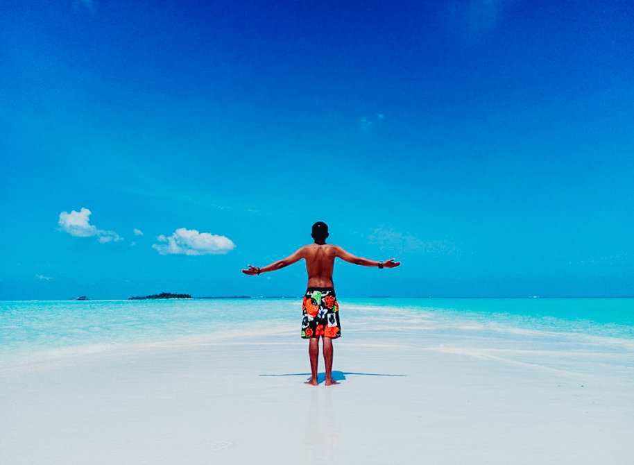 A man standing in the middle of a pristine sandbank surrounded by blue sky and bright clear blue sea in the Maldives 