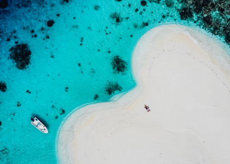 An aerial view of the sailing boat on the water stationed by a heart shaped sand bank surrounded by the bright blue clear sea 