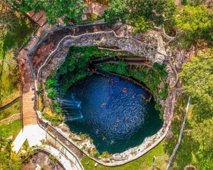 An aerial shot of a blue Cenote showing the bright blue water, luscious greenery, and people swimming in the water 