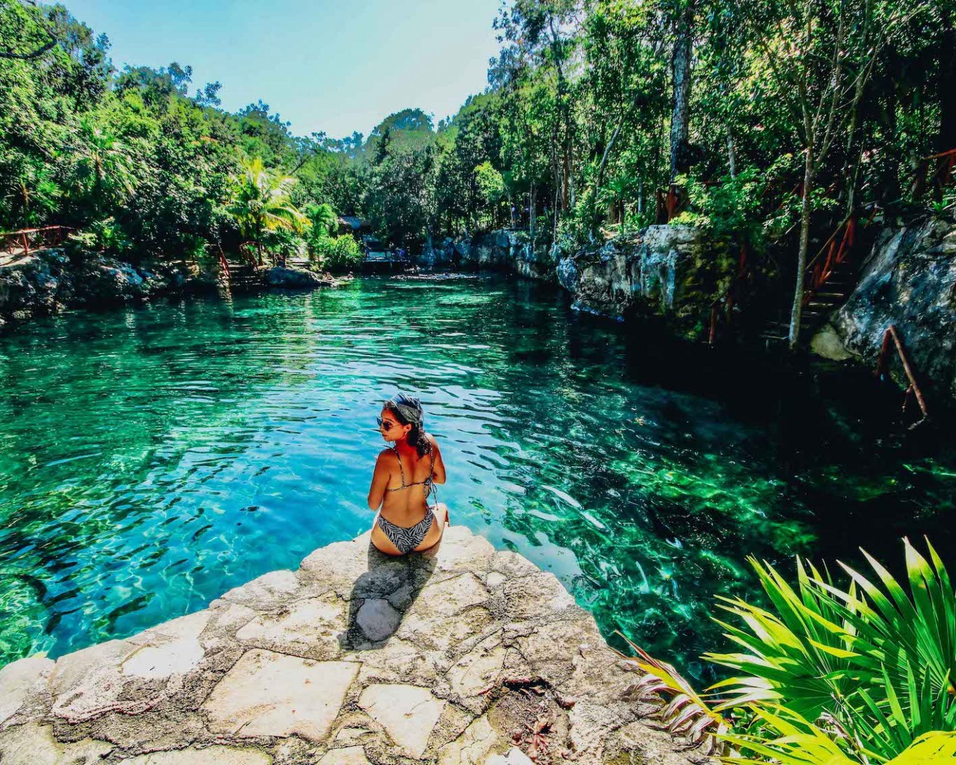 A girl in a bikini sat at the edge of a rock by bright turquoise clear water, surrounded by luscious greenery