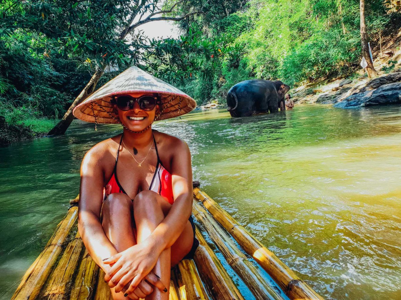 Girl sat on bamboo raft on river with asian bamboo hat on with elephant in the background 