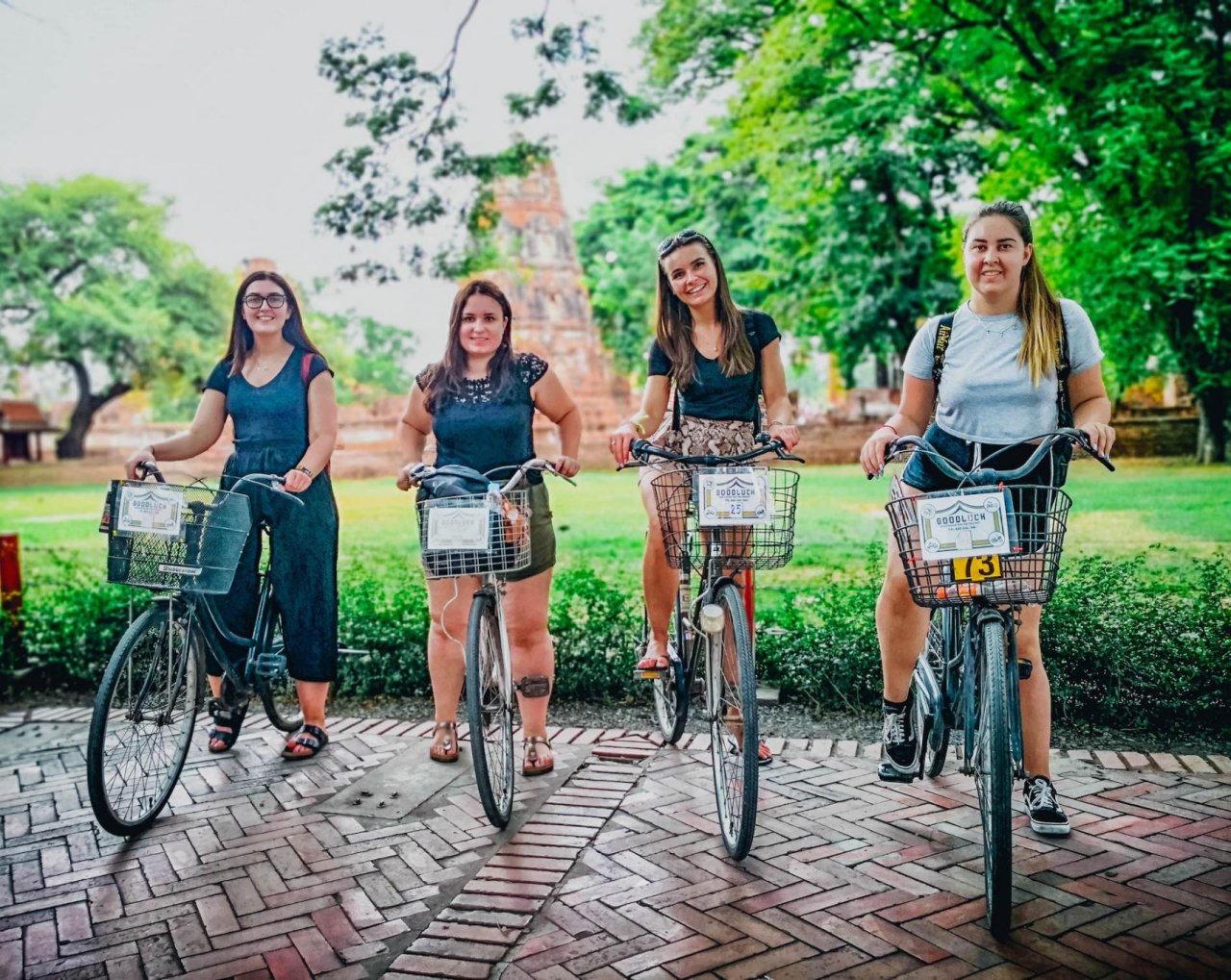 Four girls sitting on bikes with basket on the front with temples faintly showing in the background and green trees behind them