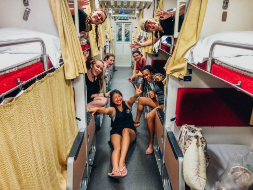 Group hanging out from bunk beds on an overnight train in Thailand with one girl sitting on the floor.