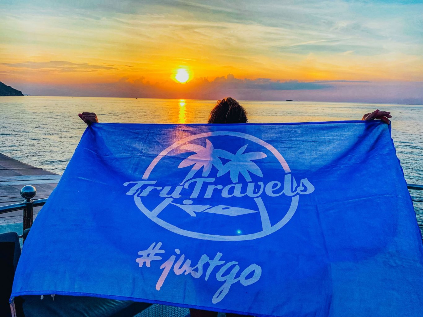 A girl holding a TruTravels flag by the beach with a stunning sunset in Thailand 