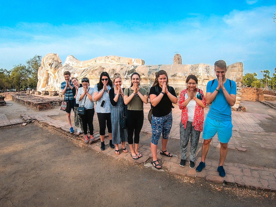 Group with hands in prayer position in front of large reclining buddha statue in Thailand